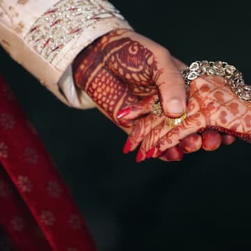 Two hands with intricate henna patterns holding each other against a dark background